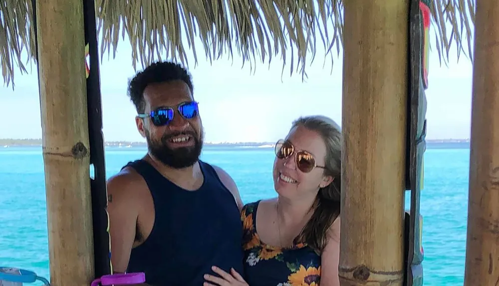 A smiling man and woman are posing for a photo under a thatched roof structure with a backdrop of a serene blue sea