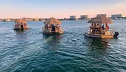 Three tiki-style boats carrying passengers are cruising on a calm body of water with a backdrop of a coastal town during sunset.
