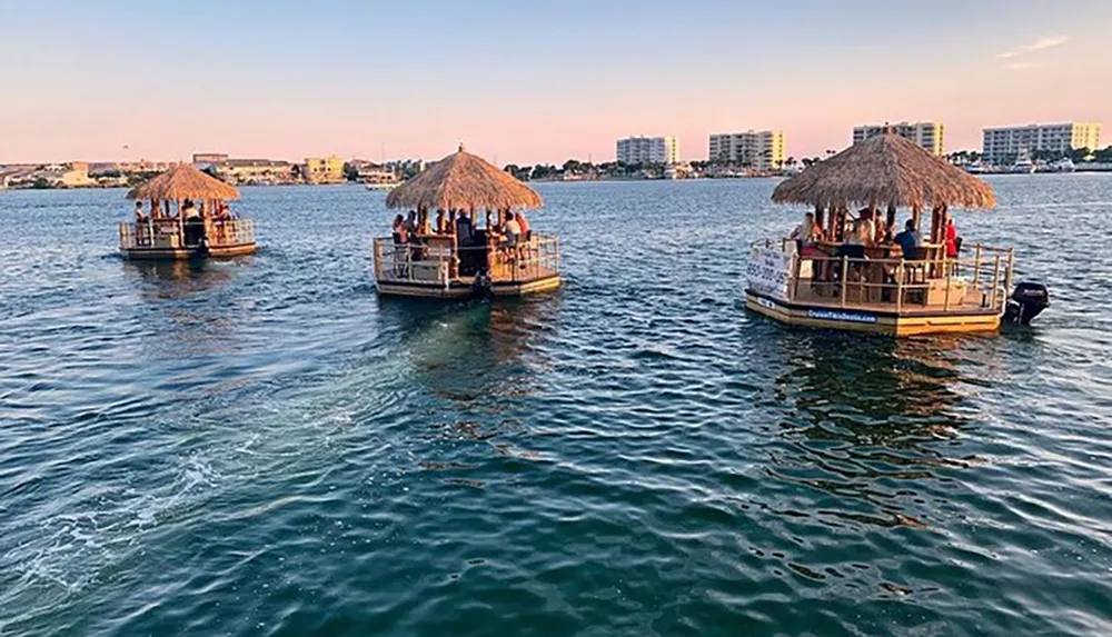 Three tiki-style boats carrying passengers are cruising on a calm body of water with a backdrop of a coastal town during sunset