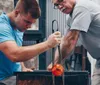 Two individuals are engaged in the intricate process of glassblowing with one person shaping a glowing molten glass object on a rod as the other observes