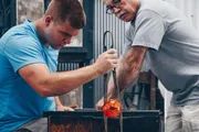 Two individuals are engaged in the intricate process of glassblowing, with one person shaping a glowing molten glass object on a rod as the other observes.