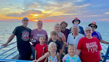 A group of people is posing for a photo on a boat at sunset with the ocean in the background.