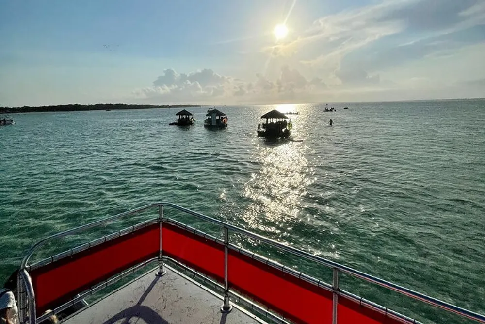 The image shows a view from the rear of a boat looking out over sunlit waters where several other boats are scattered with a setting sun low in the sky casting a glistening path across the surface of the water