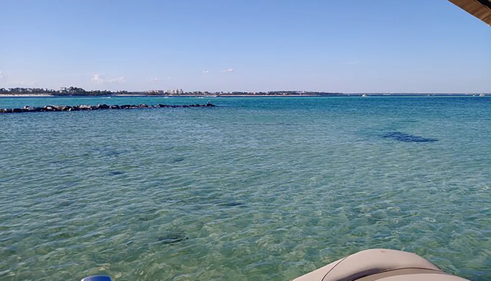 The image shows a tranquil turquoise sea viewed from the shade with a partial view of a boat in the foreground and a rocky breakwater in the distance under a clear blue sky