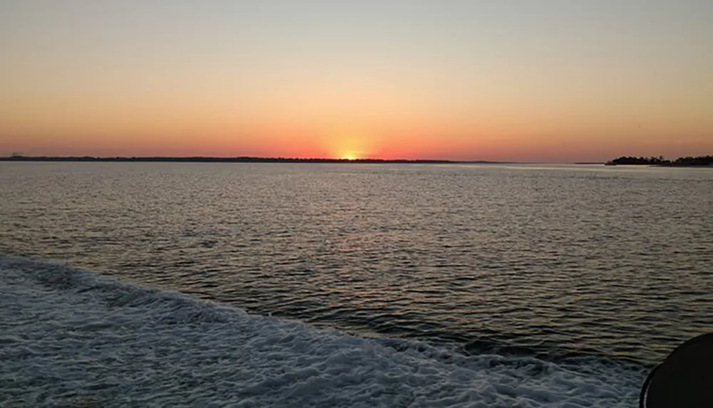 The image shows a serene sunset view over a calm body of water with a silhouette of land on the horizon and the frothy wake of a boat visible in the foreground