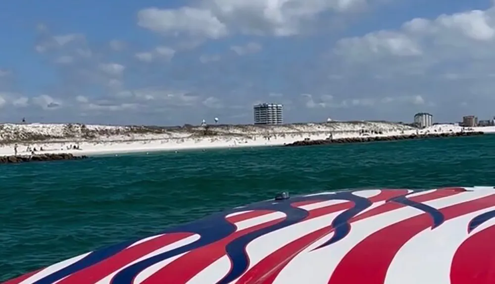 The image shows a view of a sandy beach and buildings from a boat with a red white and blue design on its bow against a backdrop of a cloudy sky