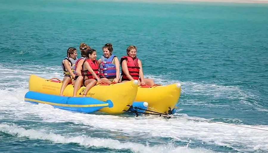 A group of people wearing life jackets are enjoying a ride on a yellow banana boat being pulled by a motorboat on a blue sea.