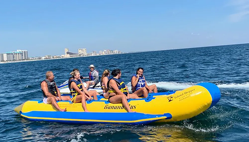 A group of people wearing life jackets are enjoying a ride on a large yellow inflatable banana boat on the sea with a coastal city skyline in the background