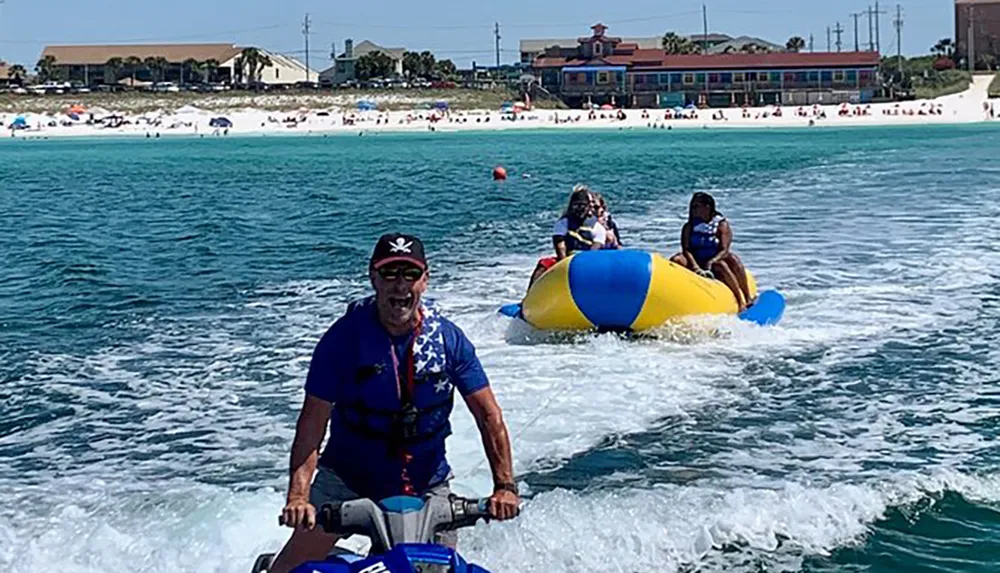 A person is operating a jet ski towing a yellow and blue inflatable with riders on a sunny day near the beach