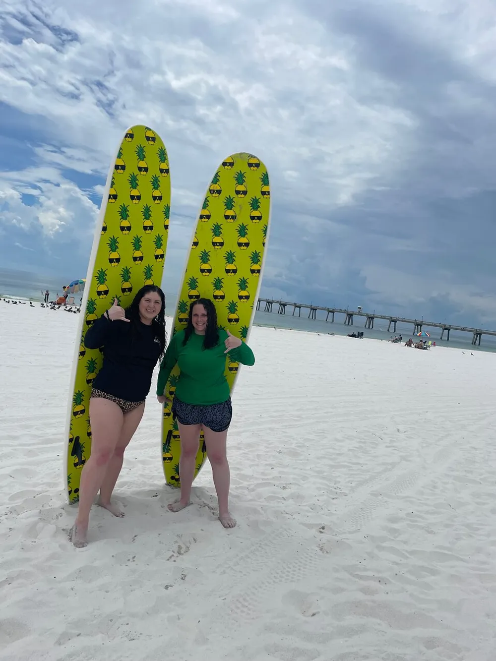 Two people are standing on a sandy beach holding bright yellow surfboards with pineapple patterns smiling for the camera