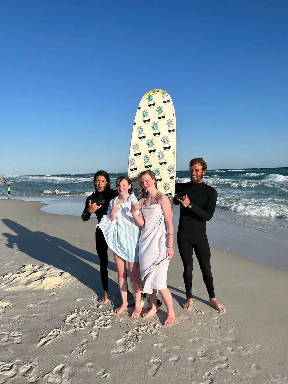 Four people are posing with a surfboard on a sunny beach two of them wrapped in towels and two wearing wetsuits all seemingly joyful