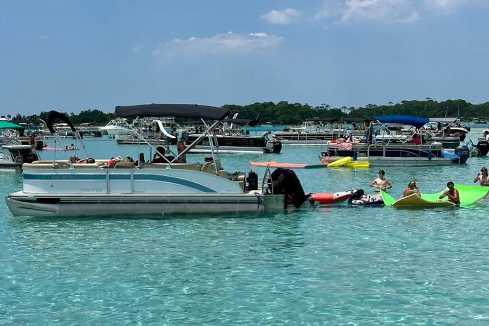 The image shows a lively gathering of people enjoying a sunny day on the water with various boats and personal watercraft floating in clear turquoise waters