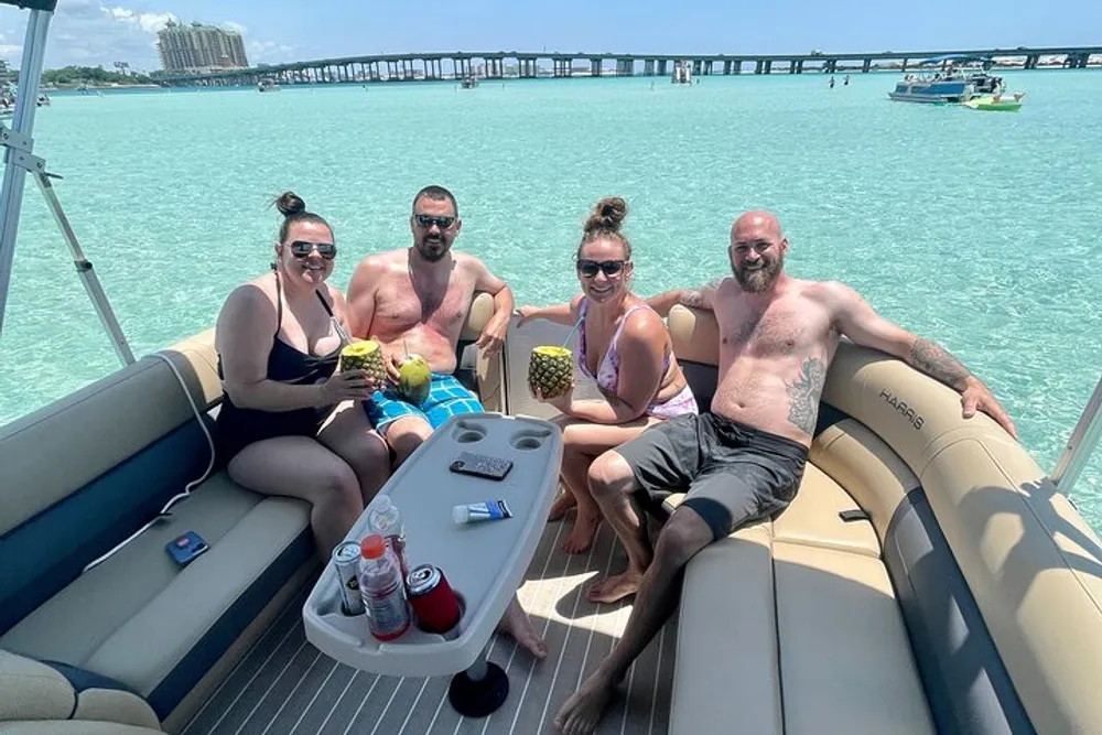Four people are enjoying a sunny day on a pontoon boat with drinks in hand surrounded by clear turquoise waters