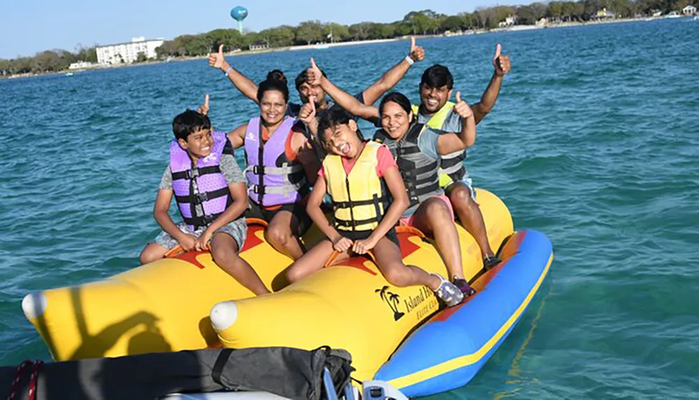 A group of people wearing life vests are enjoying a ride on a large yellow inflatable banana boat in calm blue waters with some giving thumbs-up to the camera