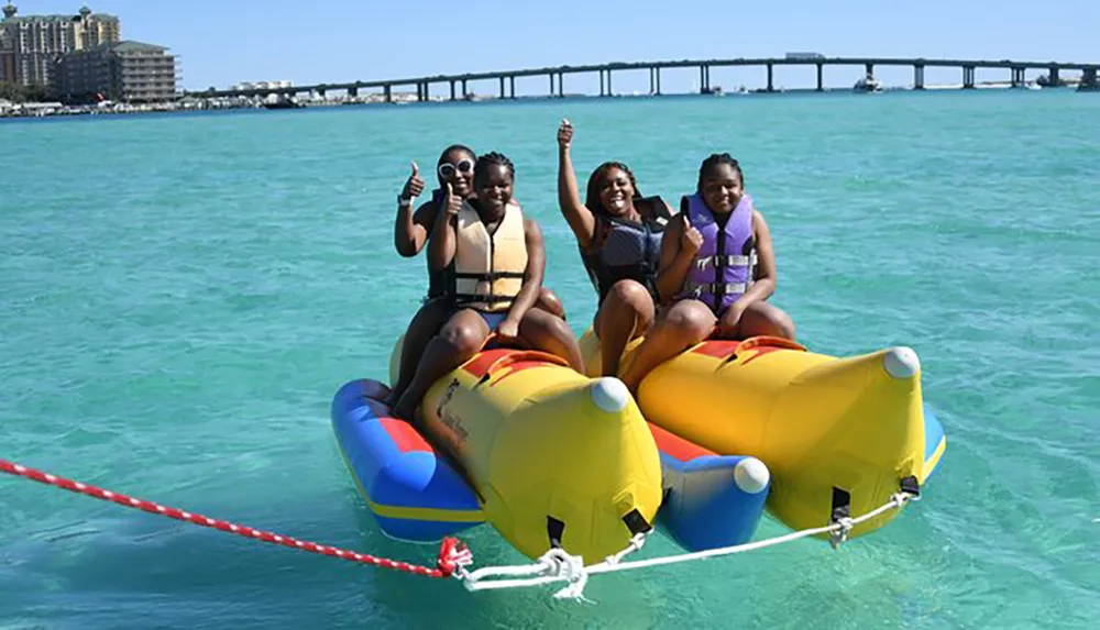 Four people are enjoying a banana boat ride on a clear turquoise sea with a bridge in the background
