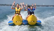 Three children are joyfully riding a banana boat on the water, with their arms raised in excitement.
