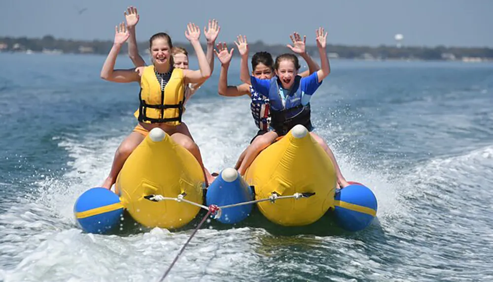 Three children are joyfully riding a banana boat on the water with their arms raised in excitement