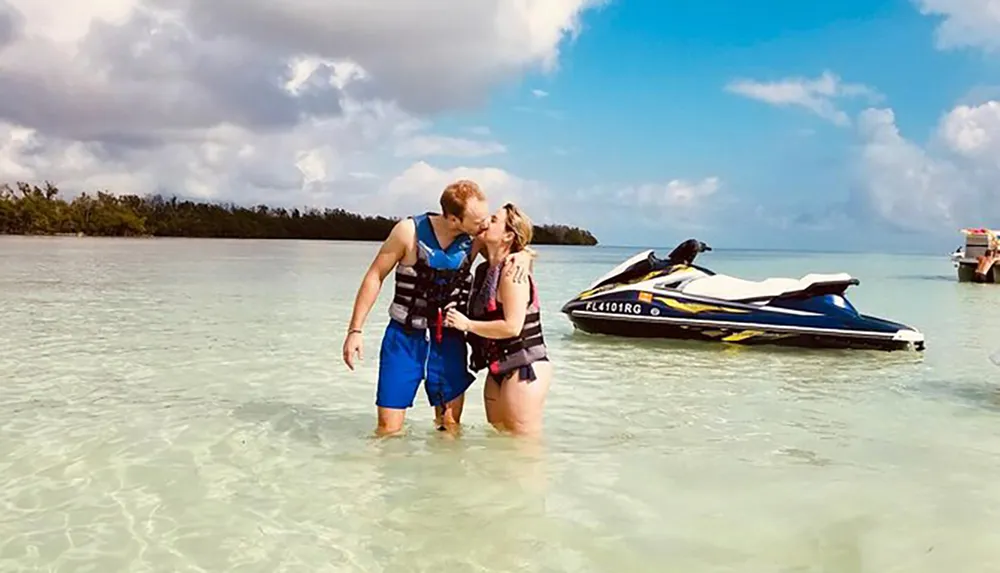 A couple is sharing a romantic moment in shallow waters near a jet ski with a backdrop of cloudy skies and distant mangroves
