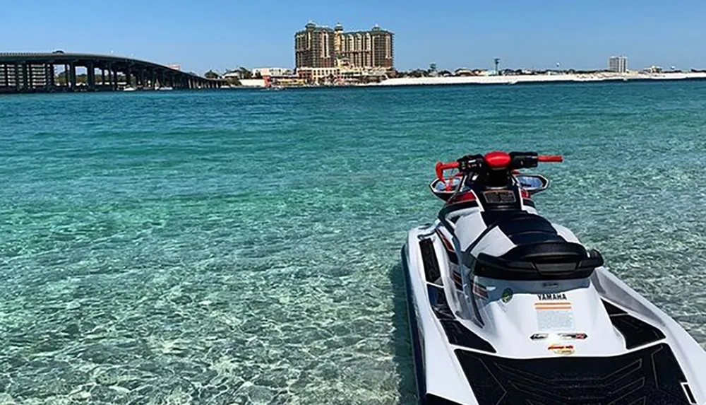 A jet ski floats on clear turquoise waters near a bridge with buildings in the background