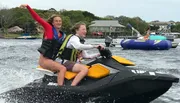 Two people are joyfully riding a jet ski on a body of water, with one person driving and the other raising her arm in excitement.