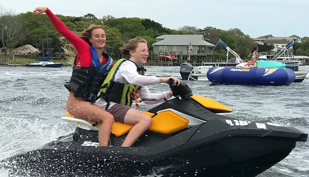 Two people are joyfully riding a jet ski on a body of water with one person driving and the other raising her arm in excitement
