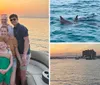 A group of six people is smiling and posing for a photo on a boat with a beautiful sunset over the water in the background