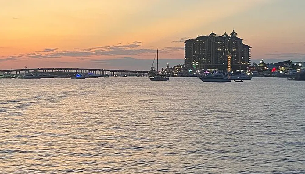 The image captures a serene sunset over a body of water with boats and a distant building skyline against a dusky sky