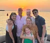 A group of six people is smiling and posing for a photo on a boat with a beautiful sunset over the water in the background