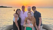 A group of six people is smiling and posing for a photo on a boat with a beautiful sunset over the water in the background.