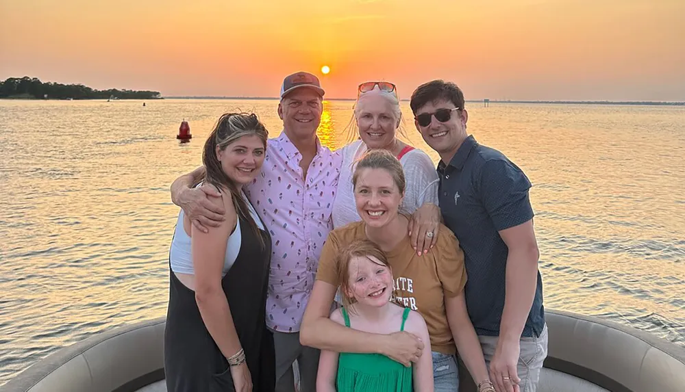 A group of six people is smiling and posing for a photo on a boat with a beautiful sunset over the water in the background