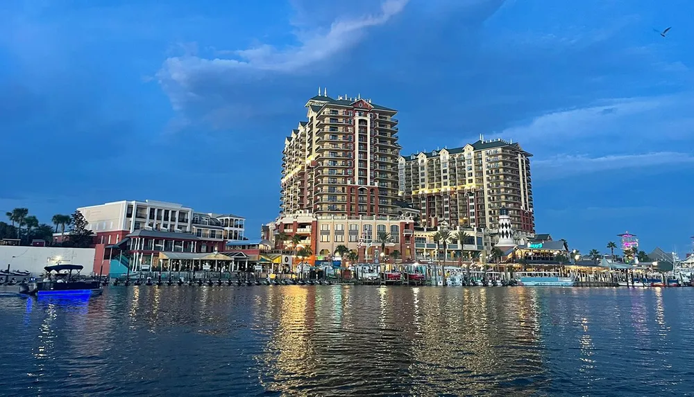 The image shows a waterfront scene at dusk with an array of buildings a calm body of water reflecting lights and a few boats docked along the pier under a sky with scattered clouds