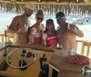 Four friends are smiling and posing for a photo at a beach bar holding drinks with a view of the water and boats in the background