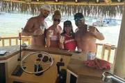 Four friends are smiling and posing for a photo at a beach bar, holding drinks, with a view of the water and boats in the background.