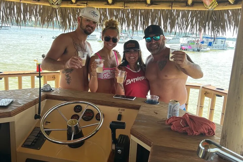 Four friends are smiling and posing for a photo at a beach bar holding drinks with a view of the water and boats in the background