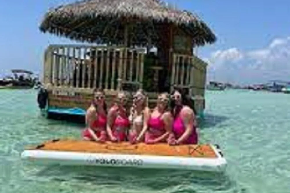 A group of individuals is enjoying time together on a floating mat in clear blue water near a thatched-roof structure