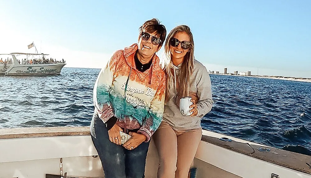 Two people are smiling and posing for a photo on a boat with the sea in the background and another boat passing by