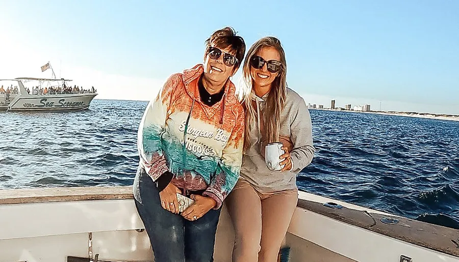Two people are smiling and posing for a photo on a boat with the sea in the background and another boat passing by.