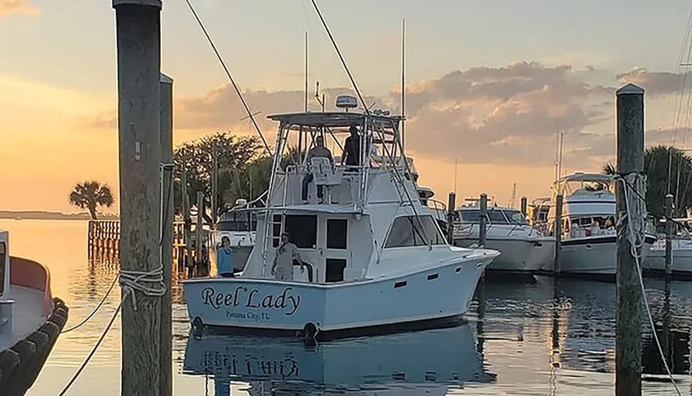 A boat named Reel Lady is docked in a calm marina at sunset with people on board and in the vicinity under a sky painted with hues of orange and peach