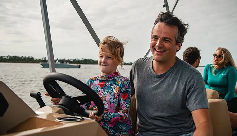A child with a big smile appears to be steering a boat with a joyful man by her side as other passengers enjoy the ride in the background