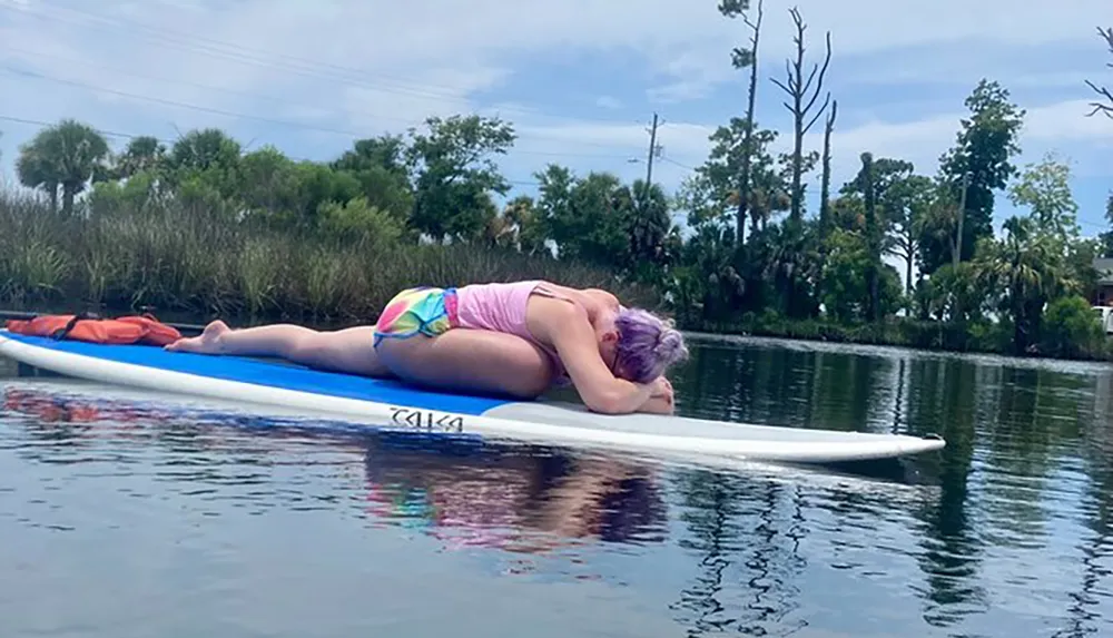 A person with purple hair is lying face down on a paddleboard in calm waters appearing to be resting or sunbathing
