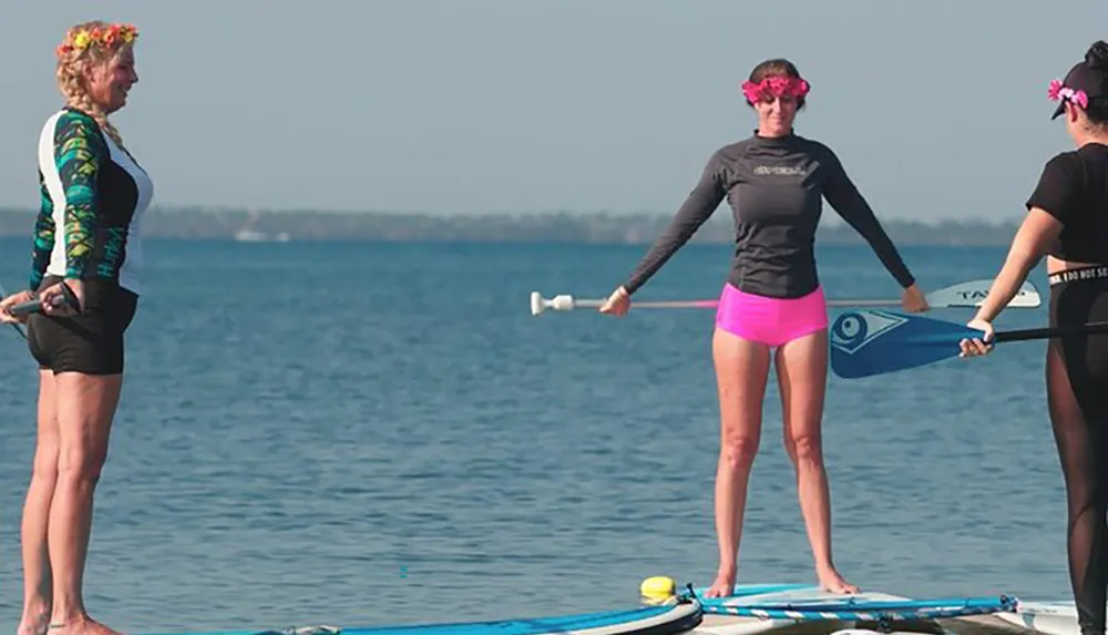 Three people are standing on paddle boards in calm waters equipped with paddles and wearing swimwear with the one in the middle wearing flower accessories in her hair