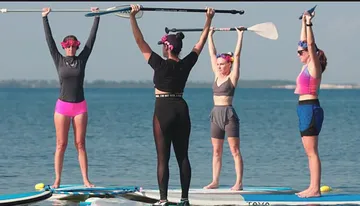 Four individuals are standing on paddleboards in a body of water, each holding a paddle overhead with a clear sky in the background.