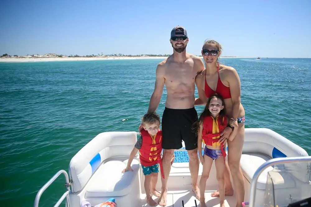 A family of four is smiling on a boat with a beach in the background and the children are wearing life jackets