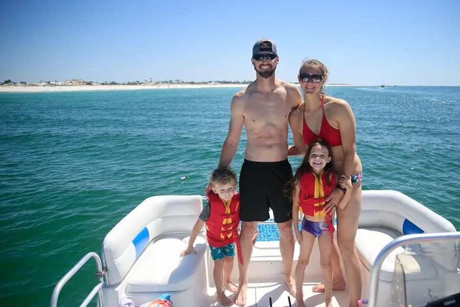A family of four is smiling on a boat with a beach in the background, and the children are wearing life jackets.