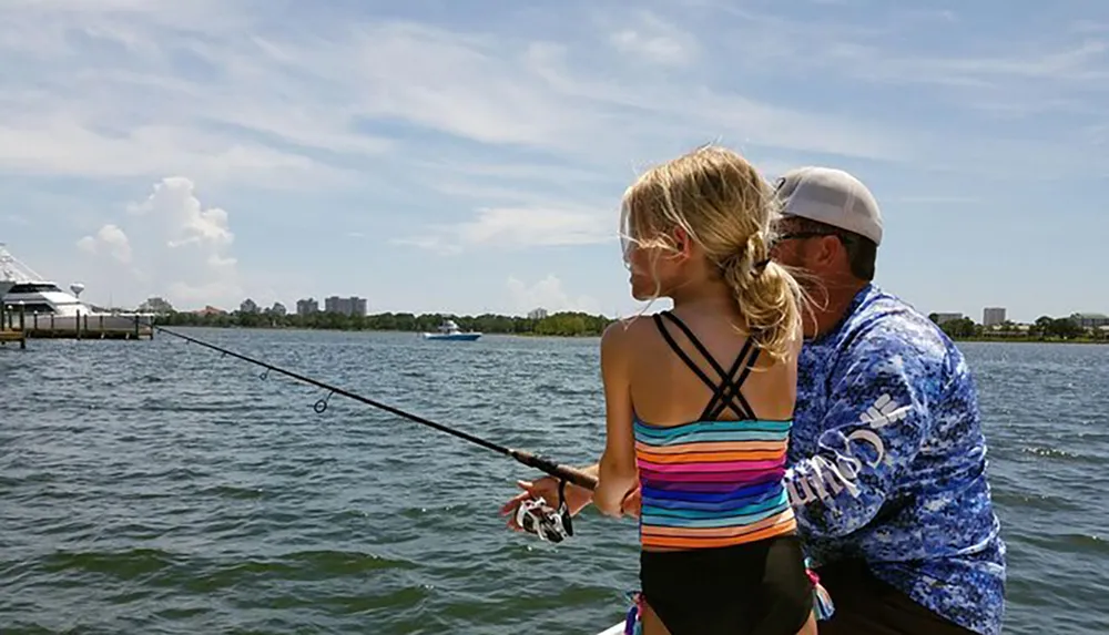 A young girl is fishing in a body of water with an adults guidance with the backdrop of a clear sky and distant buildings