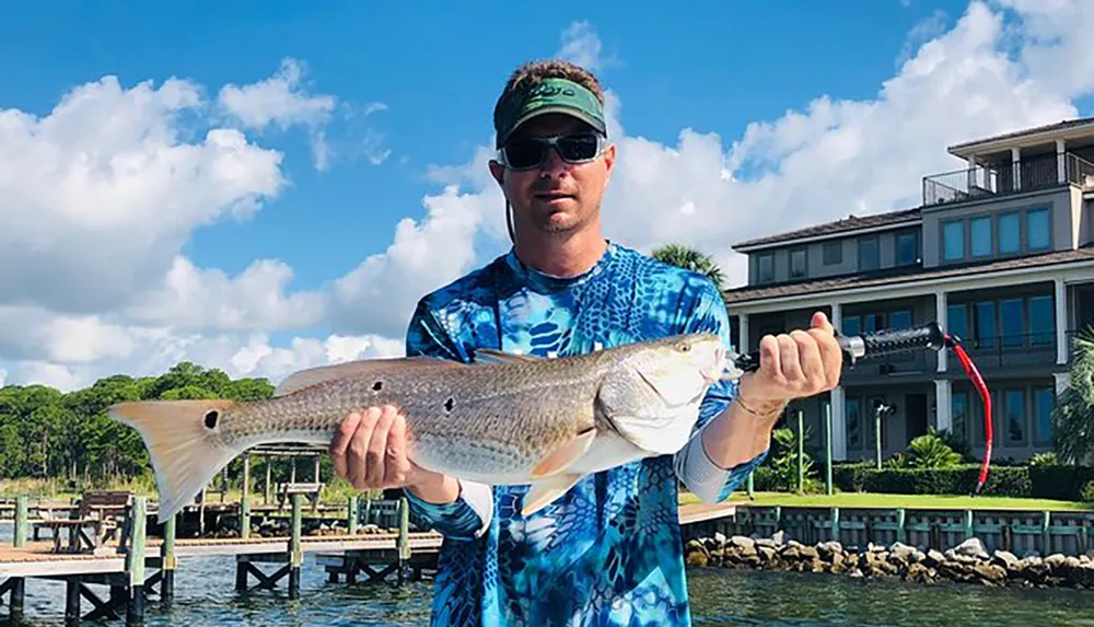 A person is proudly holding a large fish in front of a waterfront property with a clear blue sky in the background