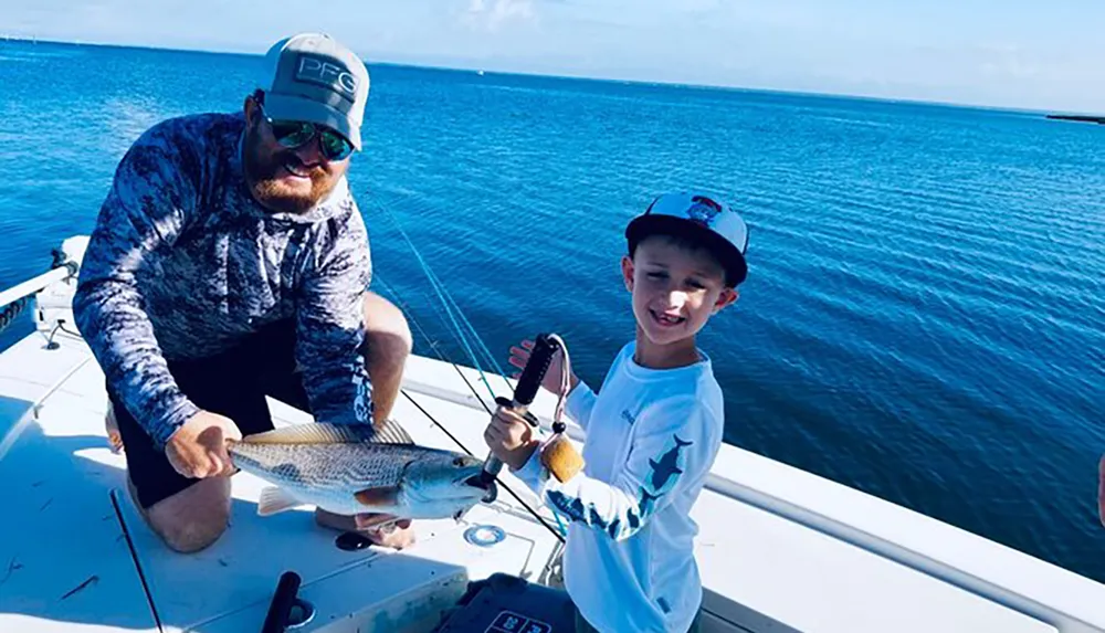 A man and a young boy are smiling on a boat showing off a fish theyve presumably caught against a backdrop of clear blue skies and calm sea waters