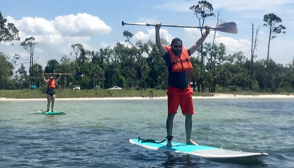 Two people are stand-up paddleboarding on a calm body of water with one person in the foreground holding their paddle overhead triumphantly