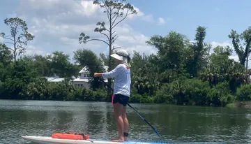 A person is paddleboarding on calm water with lush greenery in the background.