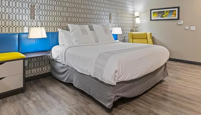 The image shows a neatly arranged modern hotel room with a large bed patterned wallpaper colorful seating and wooden flooring